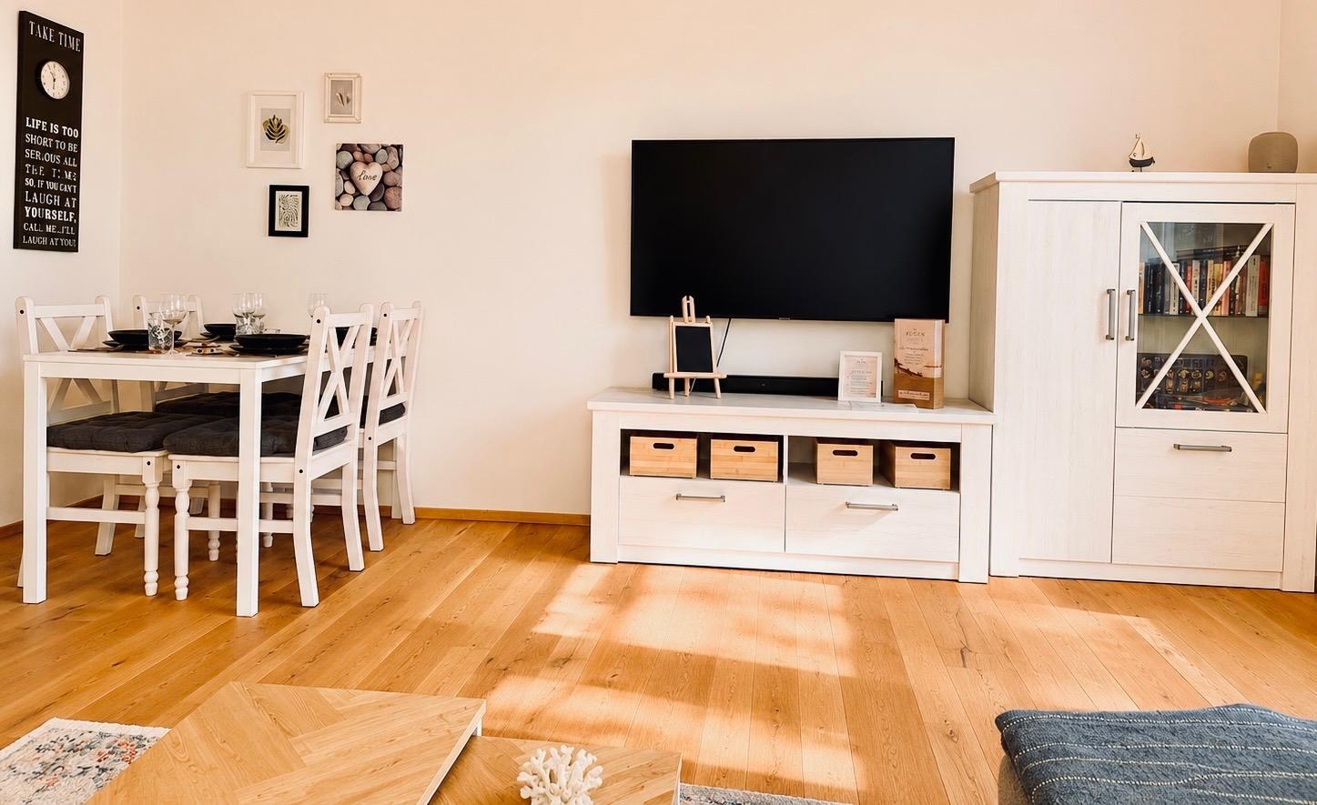 Heller Wohn- und Essbereich des DIMA Apartments mit Holzboden, Esstisch und großem Fernseher